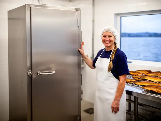 Smiling woman in a hairnet and white apron with her hand on a tall stainless smoker beside racks of smoked fish